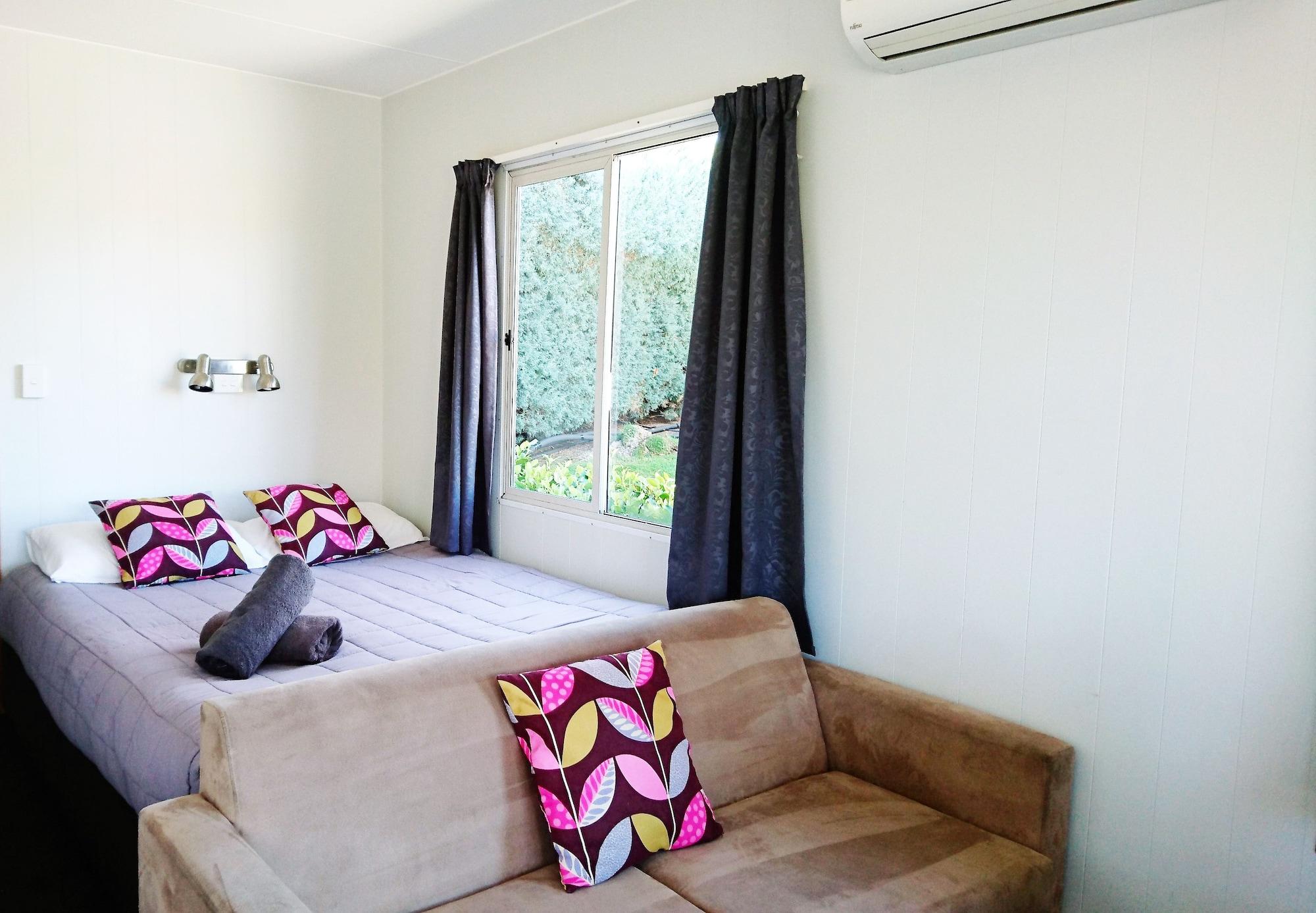 Taupo Debretts Spa Resort Екстериор снимка The photo shows a cozy room with a bed and a small sofa. The bed features two decorative pillows with colorful patterns, and there is a rolled-up towel placed on the bed. Large windows are covered by dark curtains, allowing some natural light to ente