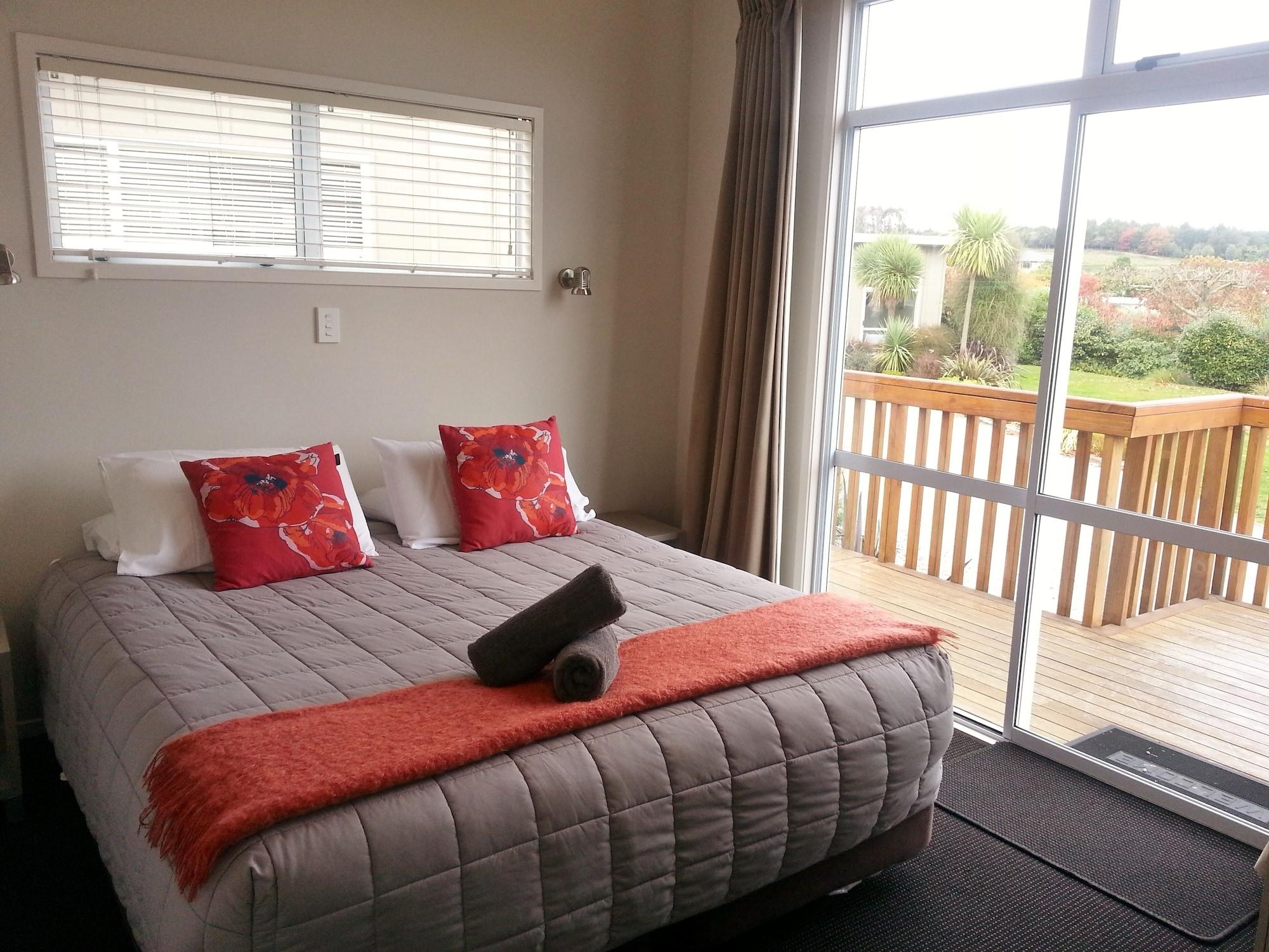 Taupo Debretts Spa Resort Екстериор снимка The photo shows a cozy bedroom with a large bed centered in the room. The bed is covered with a light gray quilt and has decorative pillows in shades of red and orange. There is a bright orange throw blanket at the foot of the bed. A rolled towel is 