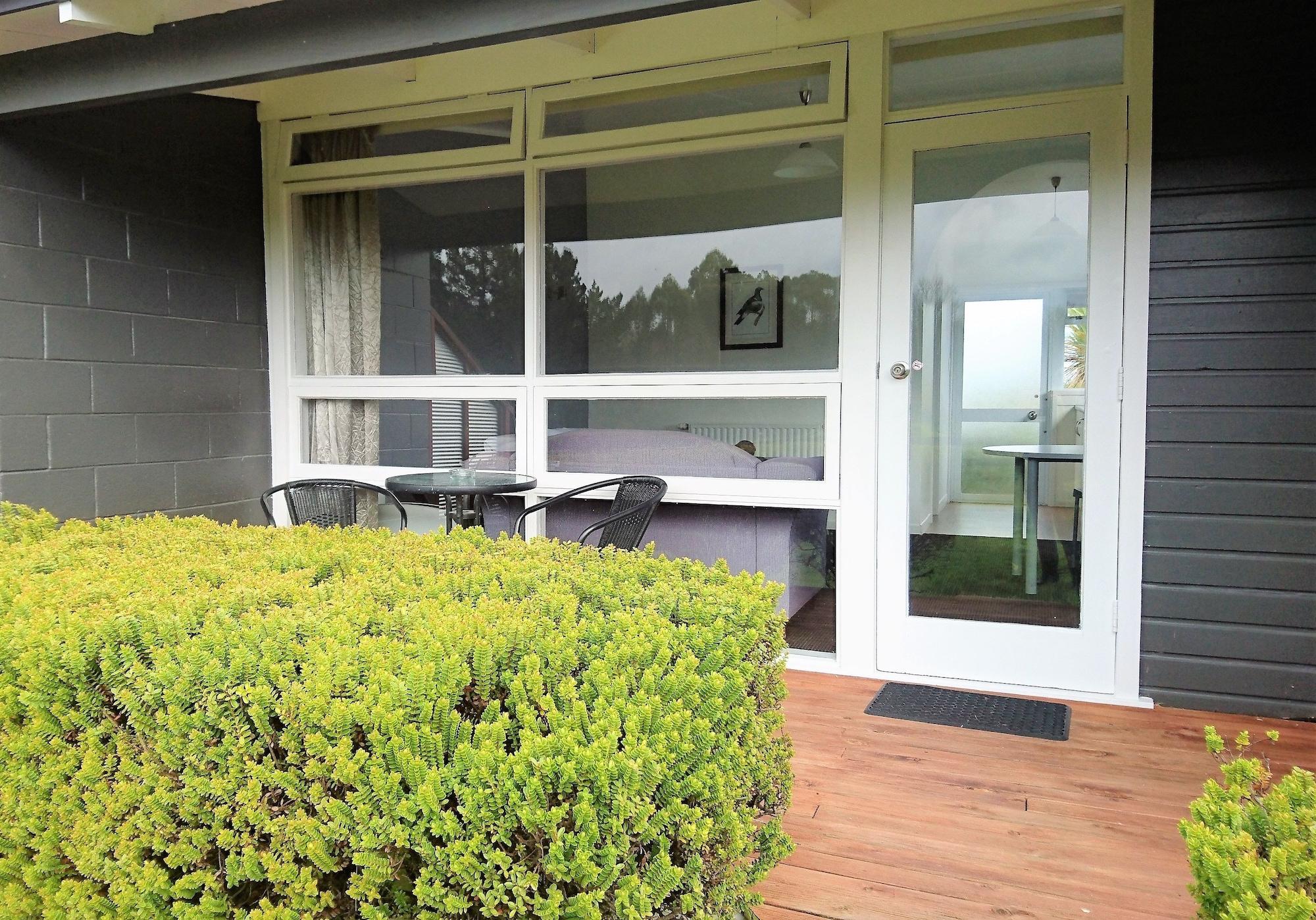 Taupo Debretts Spa Resort Екстериор снимка The photo shows a patio area in front of a building. There are green shrubs or bushes lining the foreground, creating a natural border. In the center, there is a wooden deck with a small table and two chairs, suggesting a cozy seating area. The door 