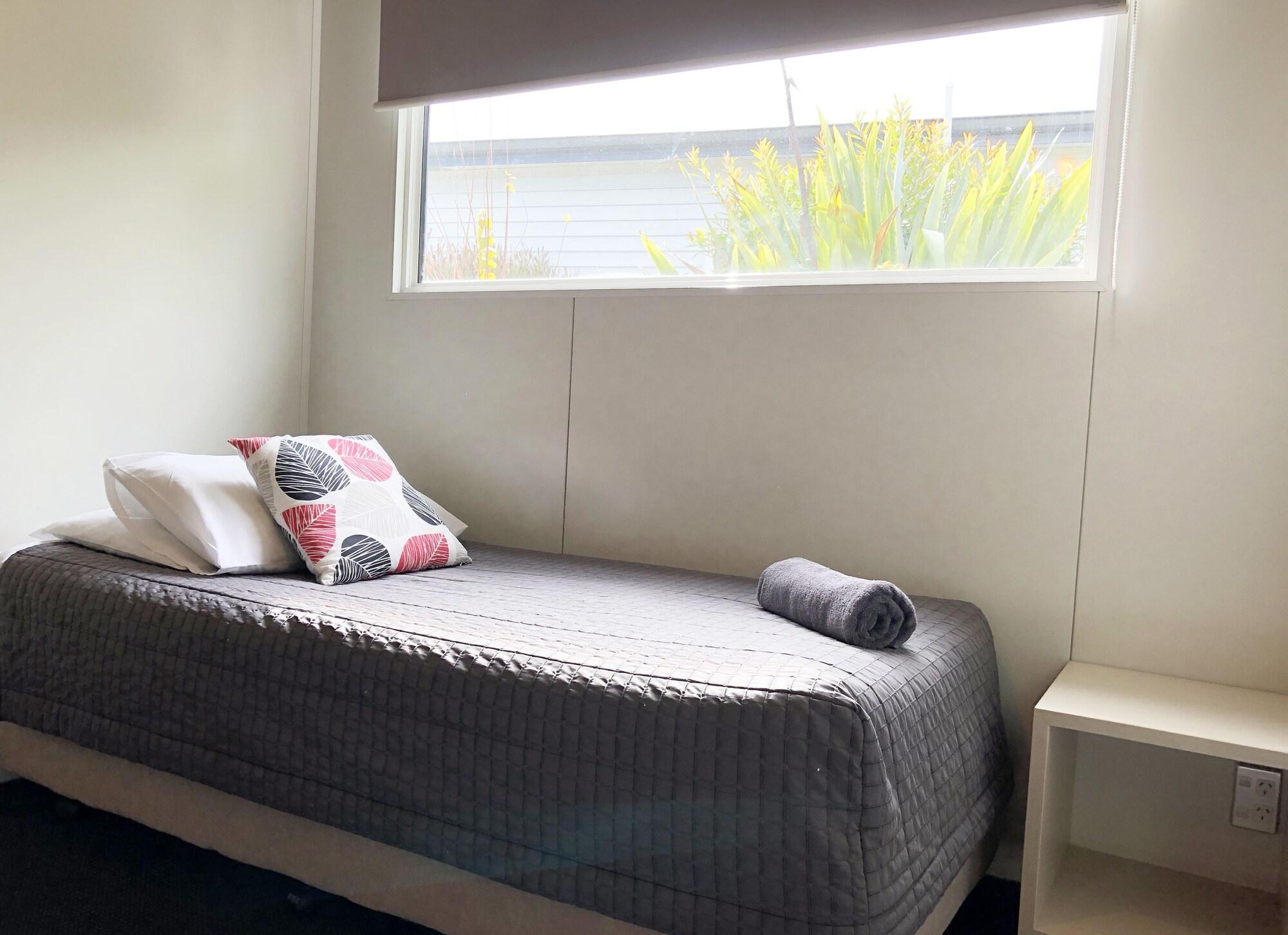 Taupo Debretts Spa Resort Екстериор снимка The photo shows a simple bedroom. It features a single bed with a gray blanket, a couple of pillows (one patterned and one plain), and a rolled-up towel placed at the foot of the bed. Next to the bed is a small white nightstand. A large window lets i