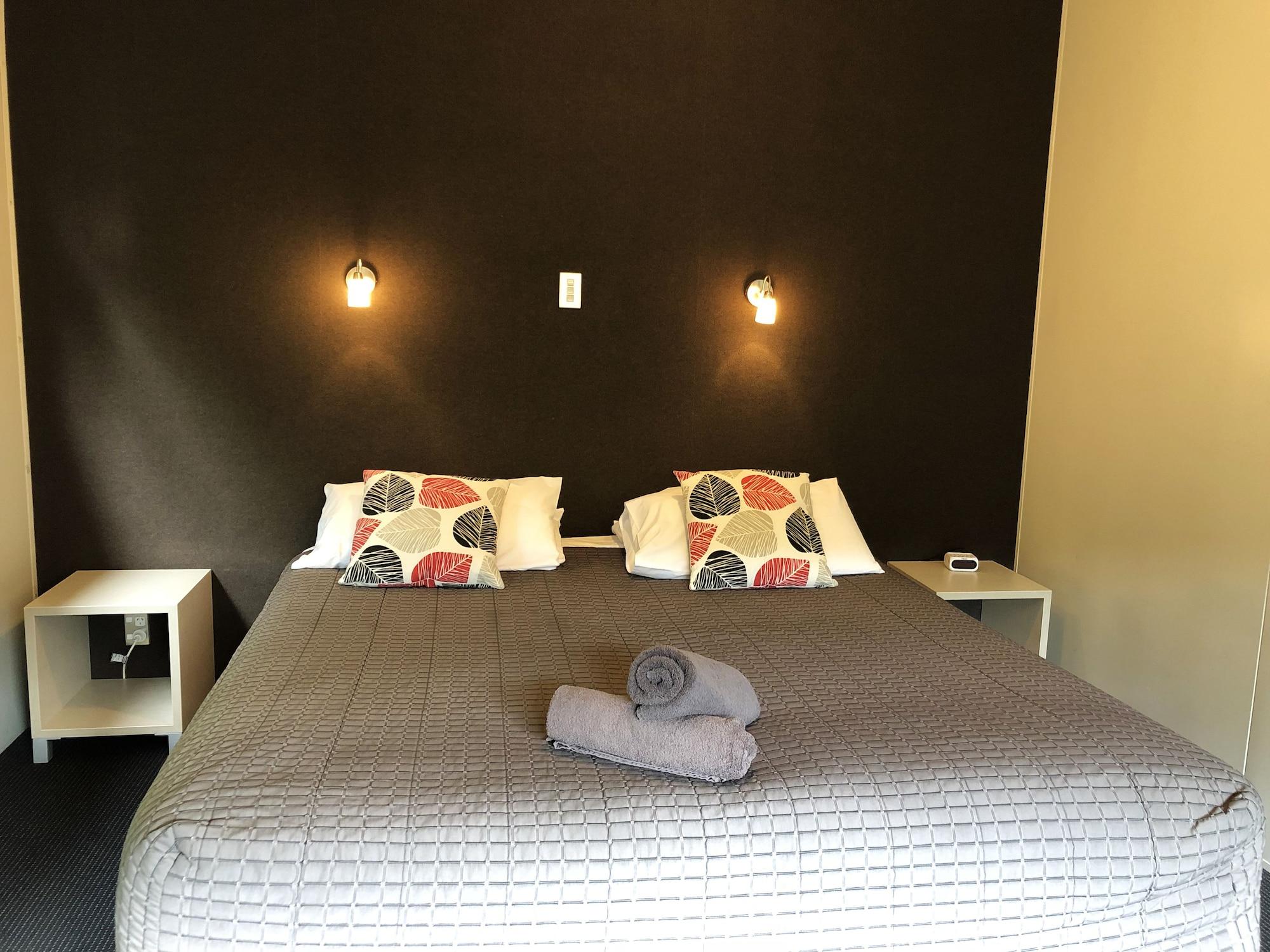 Taupo Debretts Spa Resort Екстериор снимка The photo shows a bedroom with a large bed that has a gray quilted cover and decorative pillows. The walls behind the bed are painted a dark color, and there are two small wall-mounted lamps providing light. On either side of the bed, there are small