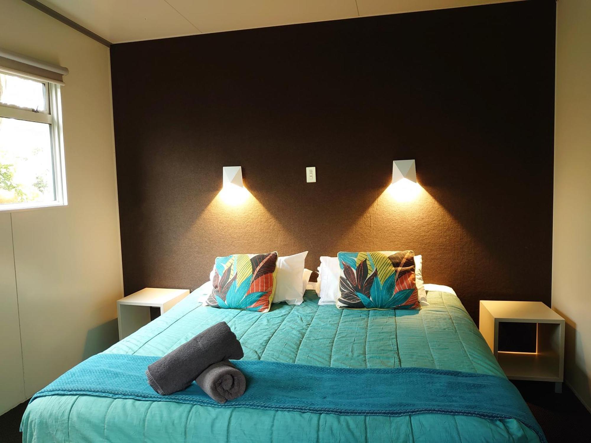 Taupo Debretts Spa Resort Екстериор снимка The image shows a bedroom with a modern and inviting layout. The focal point is a bed with a vibrant turquoise comforter, decorated with two colorful pillows featuring floral patterns. On either side of the bed, there are small nightstands with wall-