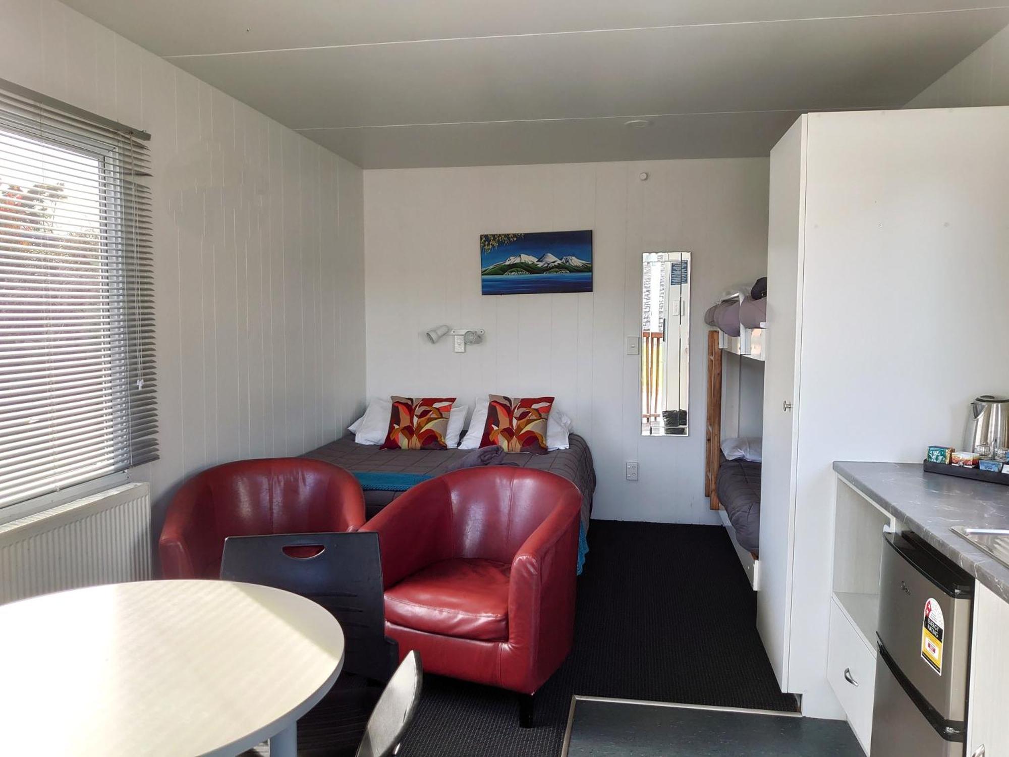 Taupo Debretts Spa Resort Екстериор снимка The photo shows the interior of a small living space, possibly a cabin or studio apartment. There are two red armchairs and a round table in the foreground. The wall opposite has a bed with colorful pillows and a small nightstand. To the side, there 