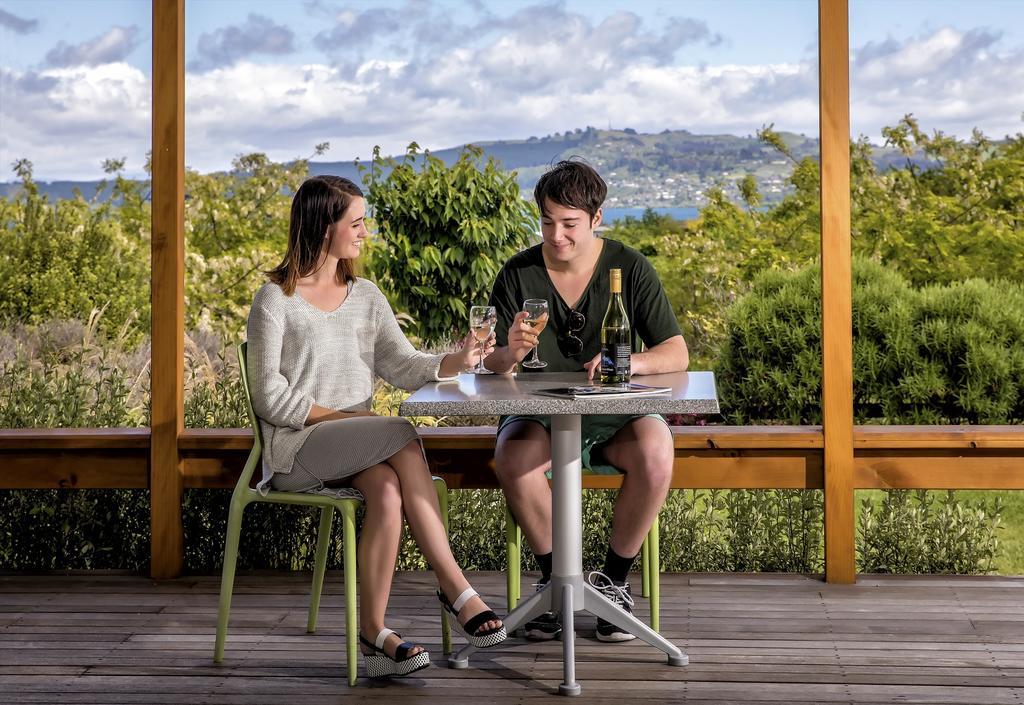 Taupo Debretts Spa Resort Екстериор снимка The photo shows a young couple sitting at a table outdoors. They are enjoying drinks, with the woman wearing a light-colored sweater and a skirt, while the man is dressed in a dark shirt and shorts. The table is set with two glasses and a bottle of w