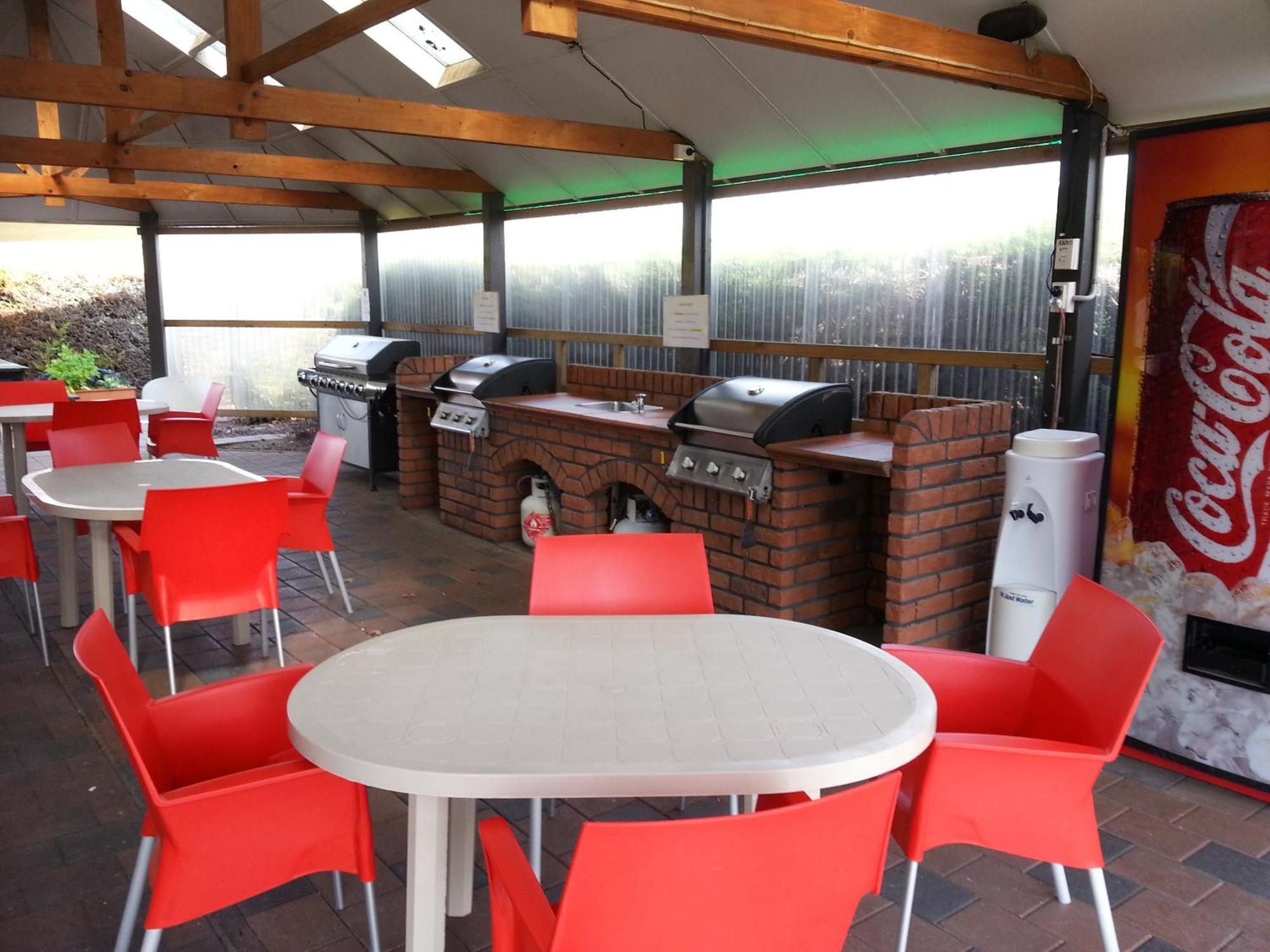 Taupo Debretts Spa Resort Екстериор снимка The photo shows an outdoor kitchen area with a covered space. There are several barbecue grills set up on a brick counter. In the foreground, there are round tables with white tops and bright red chairs arranged around them. A Coca-Cola vending machi