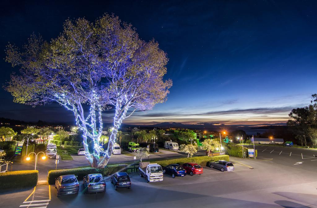 Taupo Debretts Spa Resort Екстериор снимка The photo depicts a nighttime scene of a parking lot illuminated by streetlights. In the foreground, there are several parked cars in various colors. A large tree in the center is adorned with blue lights, creating a striking visual effect. The backg
