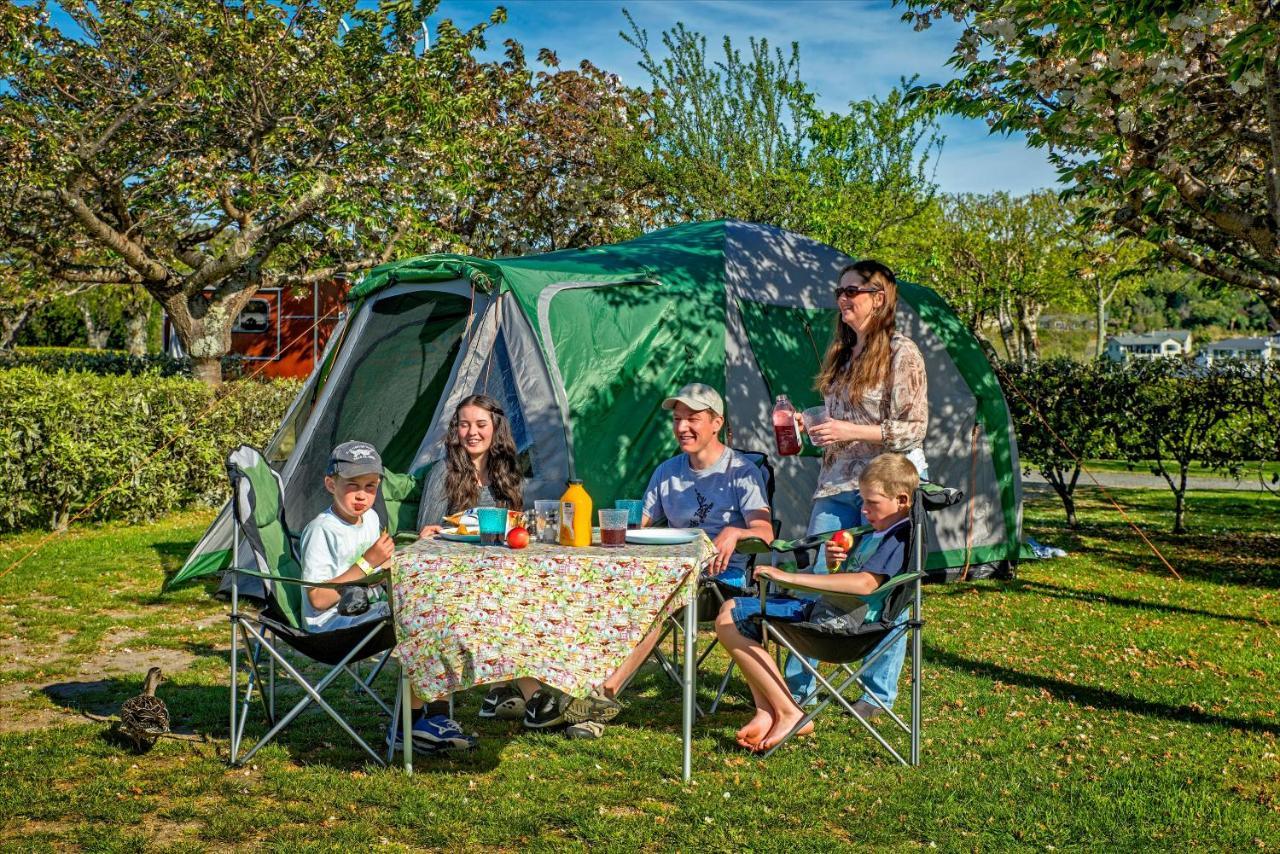 Taupo Debretts Spa Resort Екстериор снимка The photo shows a group of five people enjoying a picnic outside a green tent in a park-like setting. They are seated around a table covered with a floral tablecloth, which has various food and drink items on it. The group includes two adults and thr
