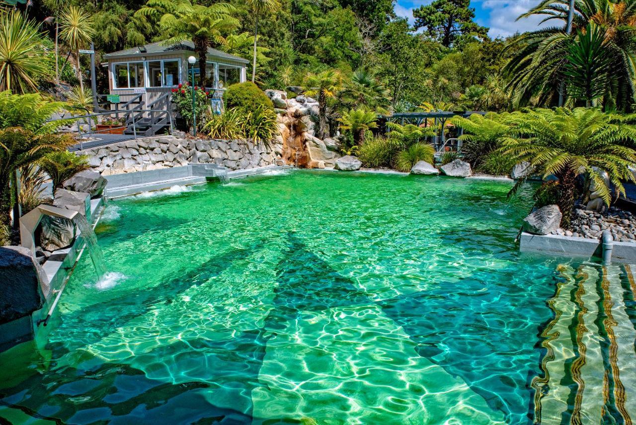 Taupo Debretts Spa Resort Екстериор снимка The photo showcases a vibrant, natural-looking pool with crystal-clear, turquoise water. Surrounding the pool are lush green plants and palm trees, creating a tropical atmosphere. In the background, there is a white building, possibly a facility or a
